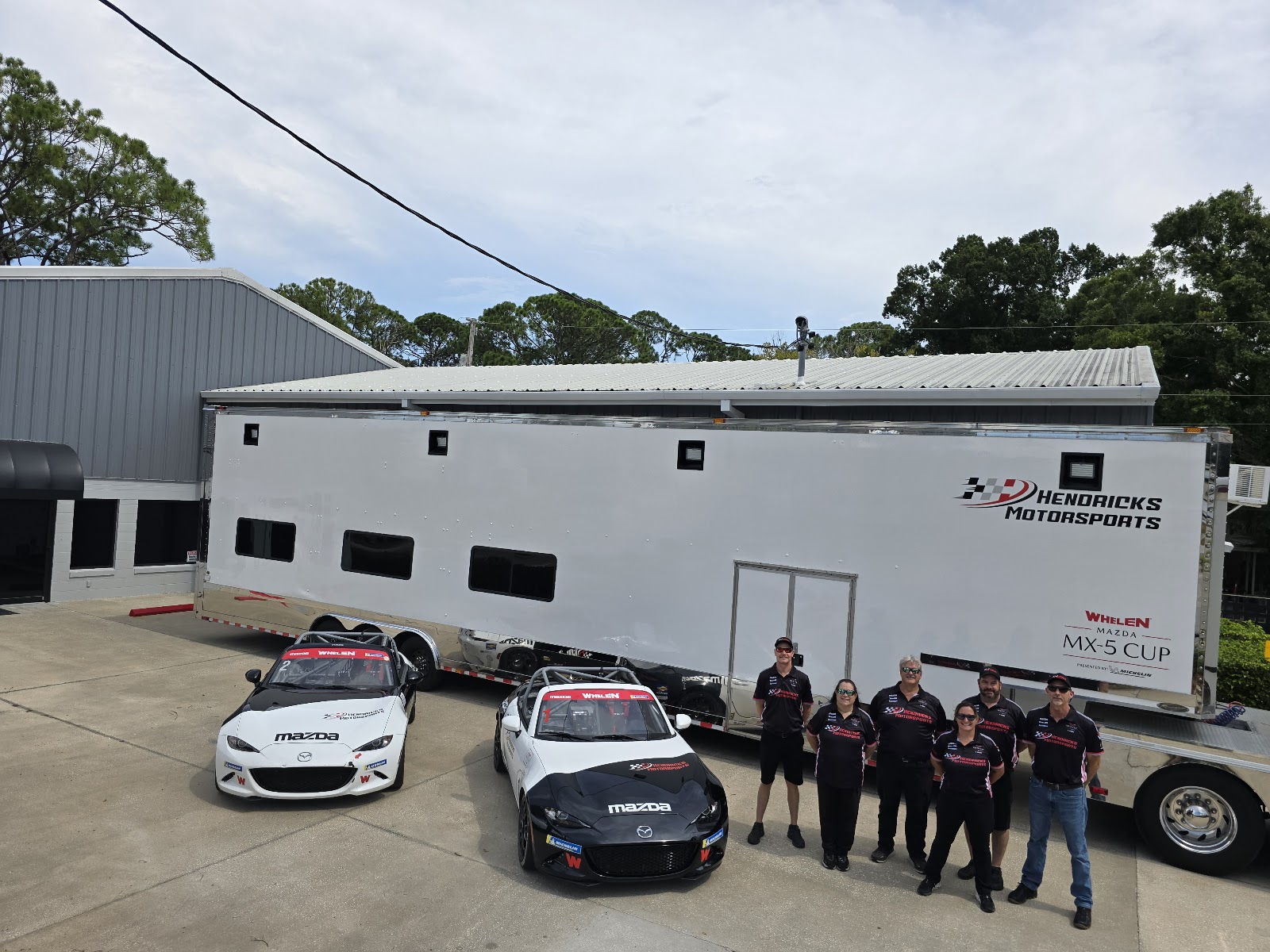 Hendricks Motorsports team in matching uniforms standing in front of the Whelen Mazda MX-5 Cup trailer at the Mason Ave shop